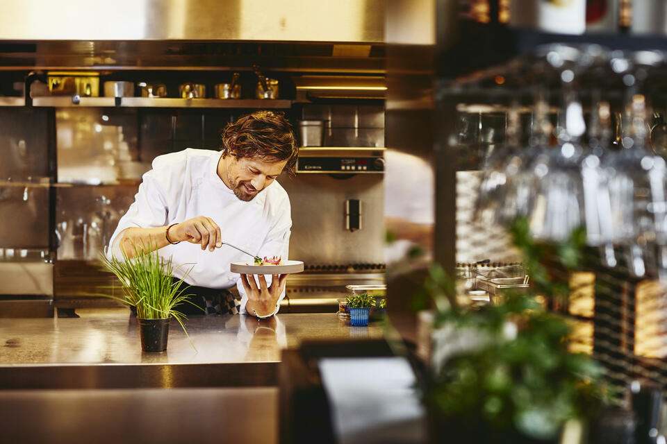 Smiling cook/chef looking down while holding a plate with food in a kitchen
